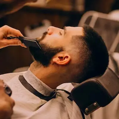 A customer sits in a barber chair with a cape as the barber trims his hair with electric clippers, against a softly blurred, welcoming barbershop interior.